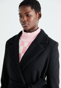 Black wool coat with a belted waist and wide lapels, worn over a ribbed pink turtleneck featuring a cream checkered pattern.