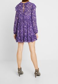 Violetti kukkakuosiset mekko pitkillä hihoilla, rypytettyjä yksityiskohtia ja avonainen selkä. Yhdistetty käärmeennahkakuosisia nilkkasandaaleja.