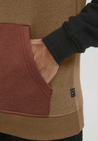 Brown and black sweatshirt with a maroon pocket, ribbed cuffs, and hem. Notable brand tag on the side, textured fabric visible.