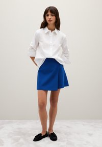 White button-up shirt with rolled sleeves, paired with a blue A-line skirt. Black slip-on shoes. Neutral background. Soft texture visible.
