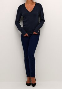 Námořnický svetr s výstřihem do V a dlouhými rukávy, zdobený zlatými knoflíky na manžetách, v kombinaci s tmavě modrými úzkými džíny a černými lodičkami.