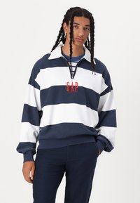 Pulóver a rayas en azul marino y blanco con cuello de cremallera, que presenta un logo rojo de "GAP" en el pecho y un corte relajado para mayor comodidad.