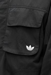 Μαύρο υφαντό μπουφάν με μεγάλη τσέπη με καπάκι και λευκό λογότυπο Adidas trefoil, με λεία υφή και λειτουργικό σχεδιασμό.