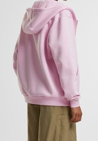 Sudadera con capucha rosa de corte relajado, puños acanalados y detalles en los bolsillos. Tejido suave y textura lisa con un acabado limpio.