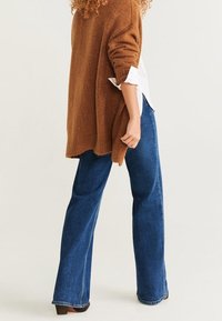 Brun oversized strikket sweater med lange ærmer, parret med blå flagrende jeans. Sweateren har en tekstureret overflade og en længere bagkant.