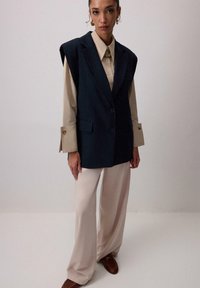 Námornícky oversized vest s dvoma gombíkmi, kombinovaný s béžovou blúzkou a širokými krémovými nohavicami. Obsahuje zlaté gombíkové detaily na rukávoch.