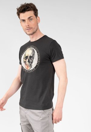 Man met kort krullend haar draagt een zwart t-shirt met schedelprint en beige cargobroek, staand tegen een effen lichte achtergrond.