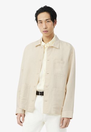 Jonge man die een beige jas met knopen draagt over een geel gestreept shirt en witte broek, staand tegen een effen lichte achtergrond.