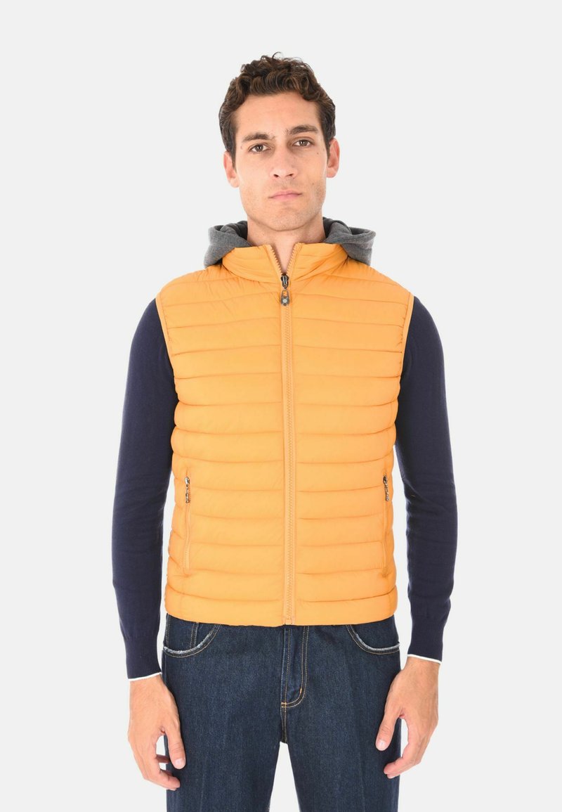 Uomo che indossa un gilet trapuntato arancione sopra una camicia a maniche lunghe blu navy e jeans blu, in piedi contro uno sfondo chiaro e uniforme.