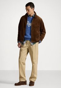 Brązowa kurtka ze sztruksu na niebieskim swetrze z białymi napisami, pod spodem koszula w kratę, beżowe spodnie i brązowe buty. Prosty design i tekstura.