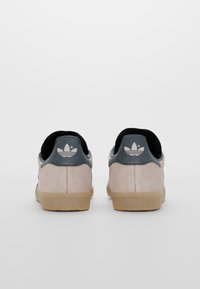 Beige mockasin sneakers med en mörkgrå hälkappa, svart inredning och sula i gummi. Vit logodetalj på hälen.