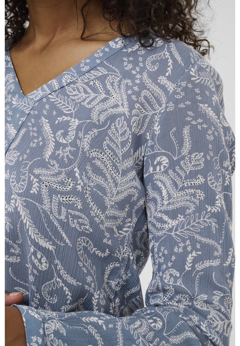 Chemise bleue à manches longues avec des motifs botaniques blancs complexes portée par une personne aux cheveux bouclés, montrée de l'épaule jusqu'au milieu du torse.