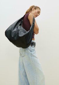 Schwarze Leder-Hobo-Tasche mit einem lässigen Design, ausgestattet mit einem Reißverschluss und einer glatten Textur. Modell trägt lässige hellblaue Jeans.