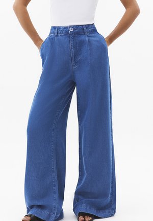 Blå jeans med vide ben båret med hænderne i lommerne, kombineret med sorte åbne sko og en hvid ærmeløs top.