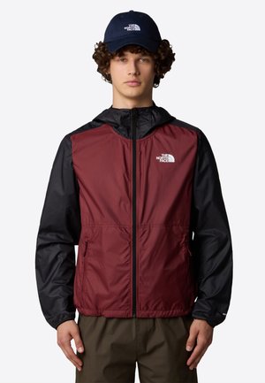 Jonge man die een bordeauxrode en zwarte North Face-jas en een marineblauwe North Face-pet draagt, staand tegen een effen achtergrond.