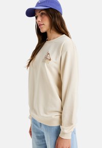 Crème-kleurige sweatshirt met lange mouwen, voorzien van ribgebreide manchetten en zoom, met een bruine logografiek op de borst. Gepromoot met een blauwe pet.