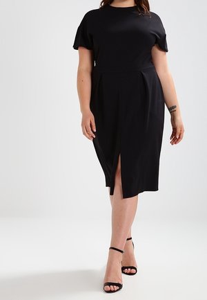 Woman wearing a knee-length black dress with short sleeves and a front slit, paired with black ankle-strap heels, standing on a plain background.