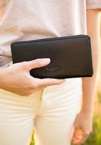 Black leather wallet held in a hand, featuring a smooth texture, zip closure, and embossed logo on the front. Rectangular shape with rounded edges.