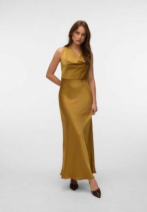 Femme aux longs cheveux bruns portant une robe en satin doré sans manches et des chaussures sombres pointues, debout devant un fond clair uni.