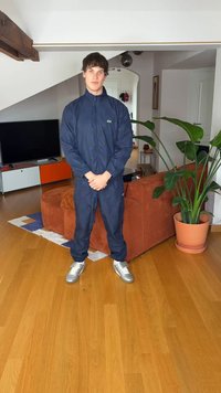 Granatowy dres z zamkiem błyskawicznym z przodu, elastycznymi mankietami i lekkim materiałem. Srebrne trampki, drewniana podłoga i doniczkowa roślina w tle.