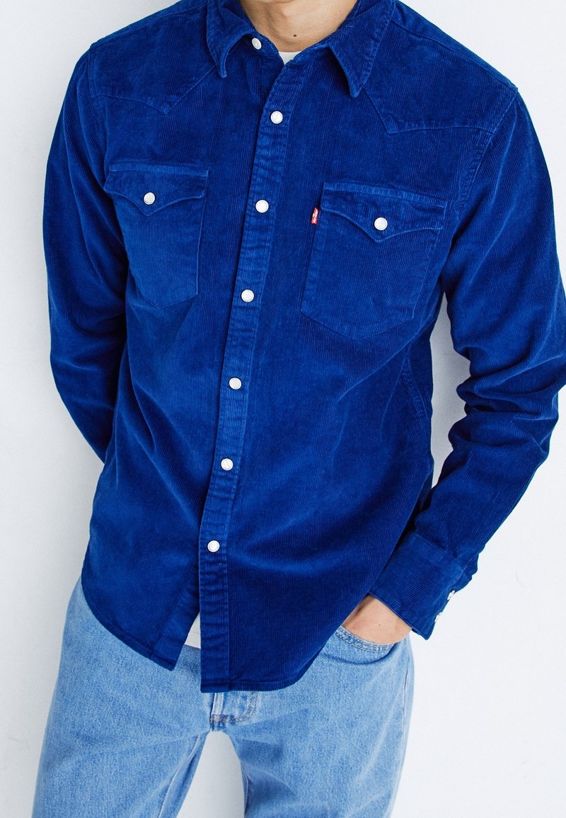 Homme portant une chemise boutonnée en velours côtelé bleu avec deux poches poitrine et un jean bleu clair, main dans la poche.