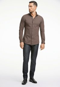 Camisa marrón de manga larga con cuello abotonado, diseño ajustado y sutil textura, combinada con jeans de mezclilla oscuros y zapatos negros.