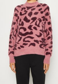 Femme portant un pull rose duveteux avec un imprimé léopard abstrait violet foncé, associé à un pantalon noir uni, debout les bras détendus.