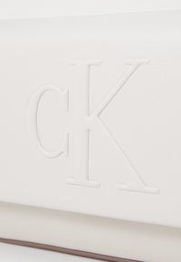 Clutch de imitación piel blanca con el logo "CK" en relieve, forma rectangular, textura suave y una costura sutil a lo largo del borde inferior.