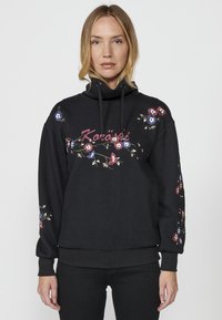 Sweatshirt noir avec texture en chevrons, broderie florale en rouge, bleu et blanc, capuche ajustable, et poignets et ourlet côtelés.