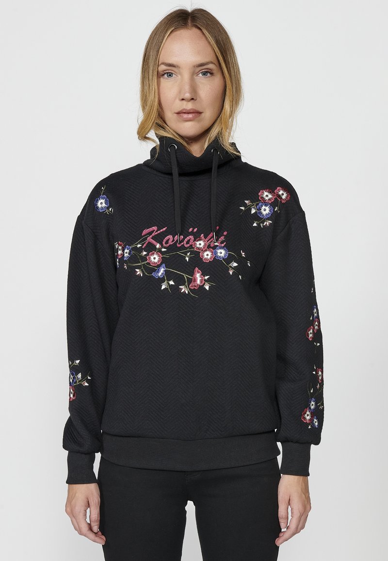 Sweatshirt noir avec texture en chevrons, broderie florale en rouge, bleu et blanc, capuche ajustable, et poignets et ourlet côtelés.