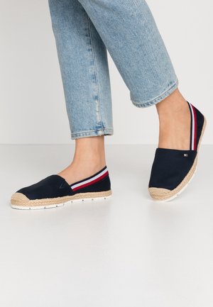 Persoon die lichtblauwe jeans en marineblauwe slip-on espadrilles draagt met rood-wit-blauw gestreepte elastische banden.