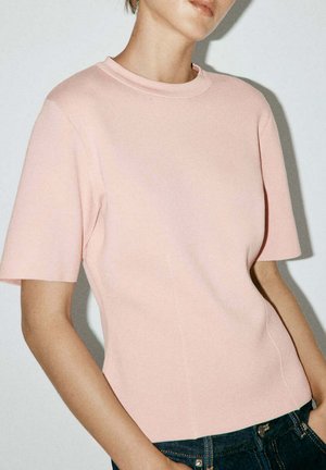 Mujer que lleva una camiseta ajustada de manga corta color rosa claro y vaqueros azul oscuro, de pie frente a un fondo liso de color claro.