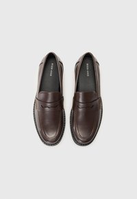 Braune Leder Slipper mit einer glatten Textur, ausgestattet mit einer geschwungenen Akzentnaht auf der Vorderseite und einer schwarzen Gummisohle.