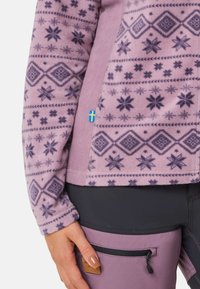 Paarse fleece top met geometrische patronen en bloemaccenten, voorzien van een klein Zweeds vlagdetail aan de zijkant en een gladde textuur.