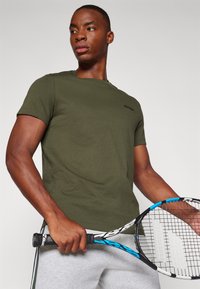 Oliven grøn t-shirt med korte ærmer lavet af blødt bomuld, med et diskret logo på brystet. Modellen holder et tennisracket med blå accenter.