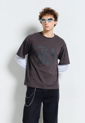 Młody mężczyzna z kręconymi włosami, ubrany w warstwowe czarno-białe długie rękawy i koszulkę z nadrukiem graficznym, czarne dżinsy, okulary przeciwsłoneczne i łańcuch jako dodatek.