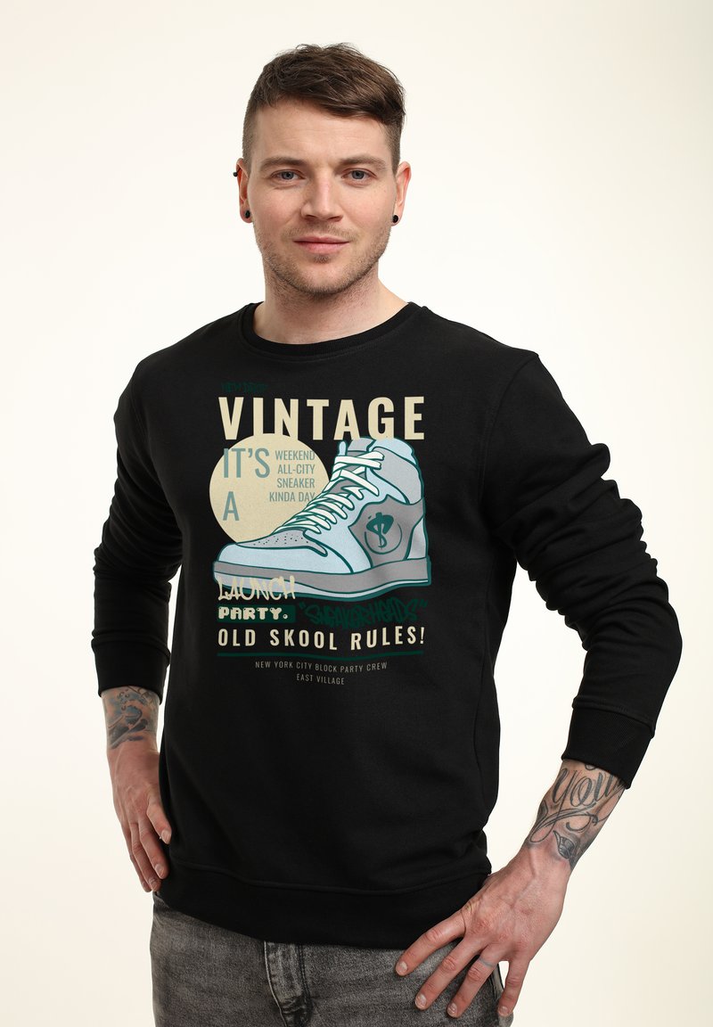 Felpa nera con maniche lunghe, caratterizzata da una stampa grafica di una scarpa da ginnastica e elementi testuali in vari colori.