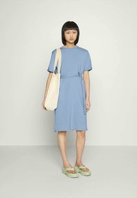 Vestido azul de comprimento até o joelho, feito de um tecido suave, com mangas curtas e uma cintura franzida. Combinado com sandálias verdes e uma bolsa tote bege.