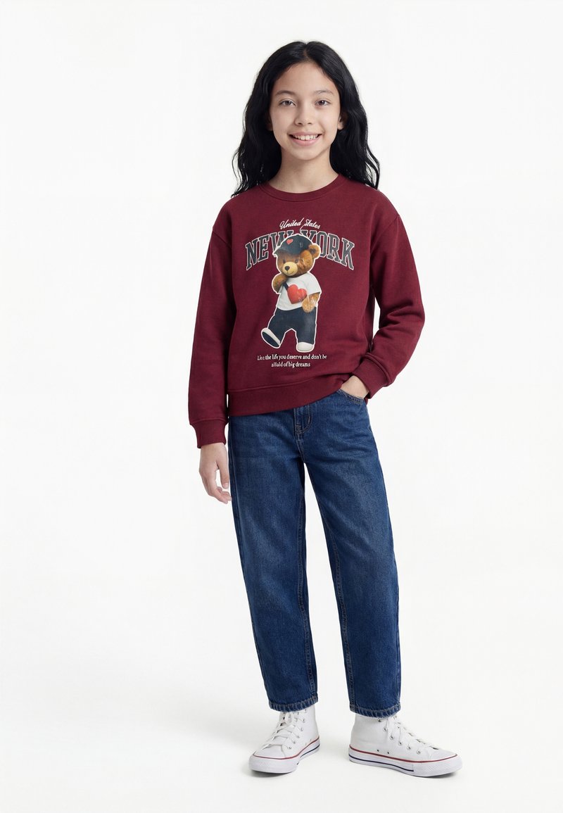 Meitene stāv, ģērbusies bordo krāsas sporta džemperī ar lāču attēlu un zilās džinsās, baltās sporta kurpēs, smaida pret baltu fonu.