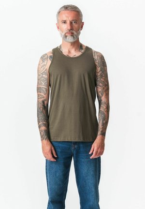 Mann med grått skjegg og tatoveringer på begge armer, iført olivengrønn tanktopp og blå jeans, stående mot hvit bakgrunn.