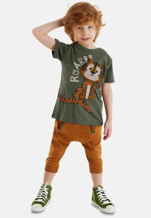 Grünes T-Shirt mit einem Cartoon-Tiger-Design und dem Text "Roarrr", kombiniert mit orangefarbenen Harem-Hosen mit Taschen und grünen High-Top-Sneakern.