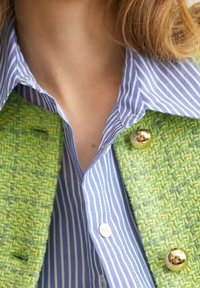 Chemise à rayures bleues et blanches avec col, sous un gilet texturé vert à boutons dorés, montrant un gros plan du décolleté et de la partie supérieure de la poitrine.