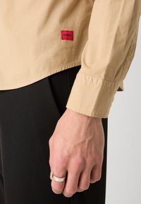 Beige long-sleeve shirt with a red "HUGO" label, textured fabric, buttoned cuff, paired with black pants and a silver ring on the hand.