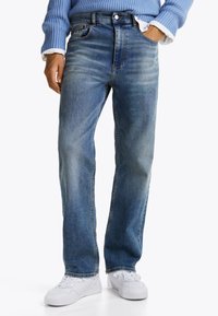 Vaqueros de denim azul claro con un diseño de pierna recta, ligero desgaste y estilo tradicional de cinco bolsillos. Combinados con zapatillas blancas.