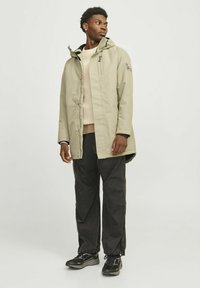 Lys beige vandtæt jakke med høj krave og hætte, lynlås lukning og sorte detaljer. Båret over en tan farvet sweater og mørke bukser.
