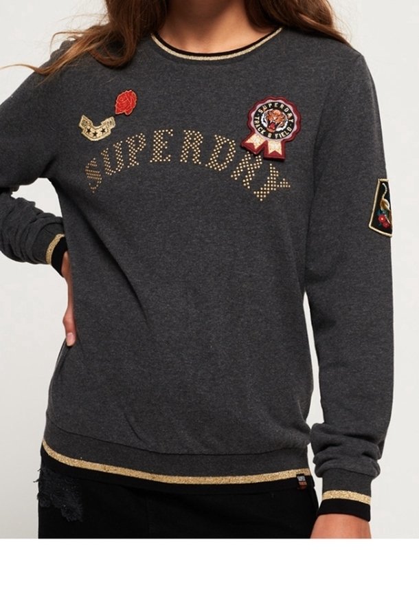 Sweat-shirt gris foncé avec le texte "SUPERDRY" en or, patches brodés incluant un emblème de tigre et des poignets et ourlet à rayures dorées.