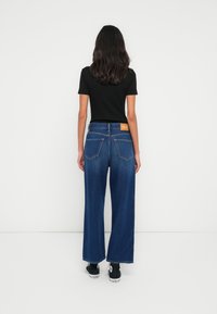 Τζιν παντελόνια με φαρδιά μπατζάκια σε σκούρο μπλε πλύσιμο, με ψηλή μέση, αντίθετες ραφές και πίσω τσέπες. Συνδυασμένα με μαύρα αθλητικά παπούτσια.