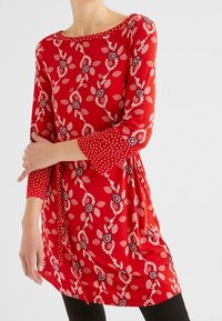 Punainen tunika, jossa on kukka- ja pilkkukuviot. Siinä on pitkät hihat ja pyöreä pääntie. Valmistettu pehmeästä, kevyestä kankaasta.