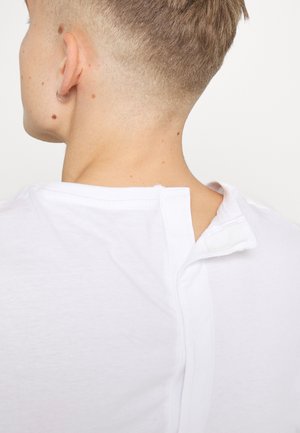 Person med kort blondt hår set bagfra iført en hvid top med en lukkedetalje ved halsudskæringen og en lille hoop-ørering i venstre øre.