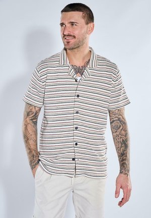 Mann mit Tätowierungen, der ein kurzärmeliges, gestreiftes Hemd mit Knöpfen und helle Hosen trägt, vor einem schlichten Hintergrund steht.